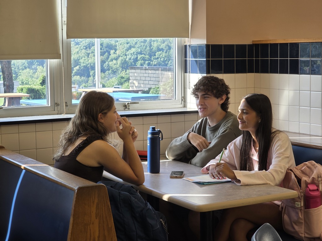 Three students sit around a table in a cafeteria, with a window behind them showing trees.