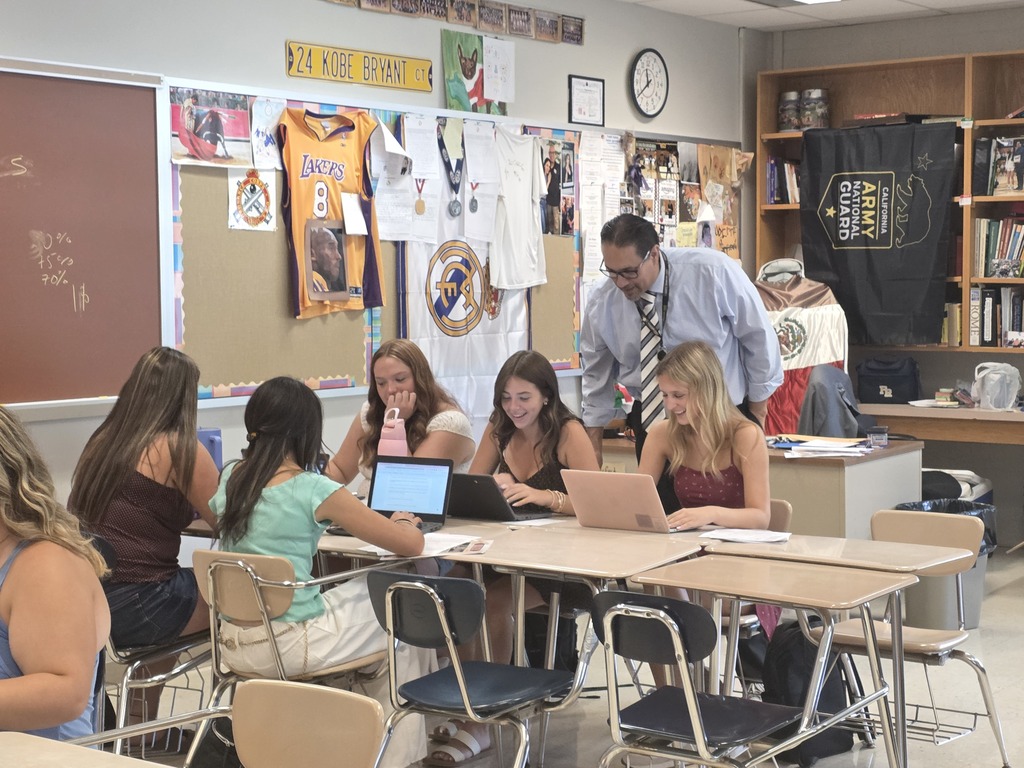 Classroom with students using laptops. Teacher stands behind them. Bulletin board and shelves in background.