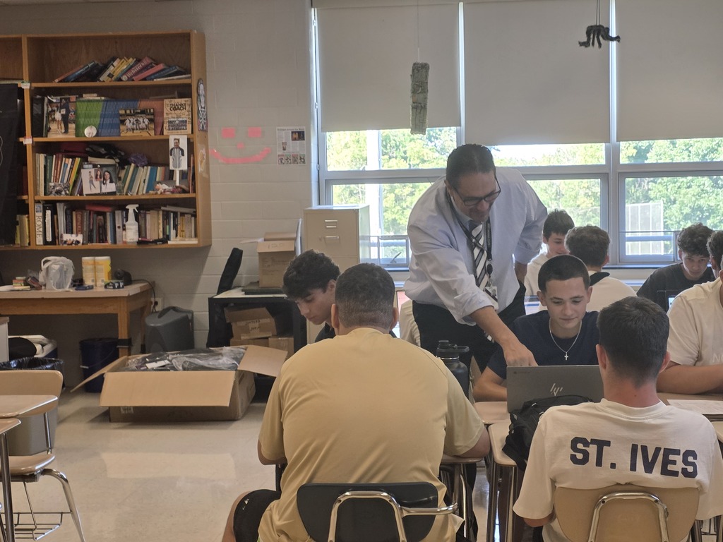 A classroom with students seated around desks; one wears a white shirt with "ST. IVES" printed on it. A teacher stands at a table with a laptop.