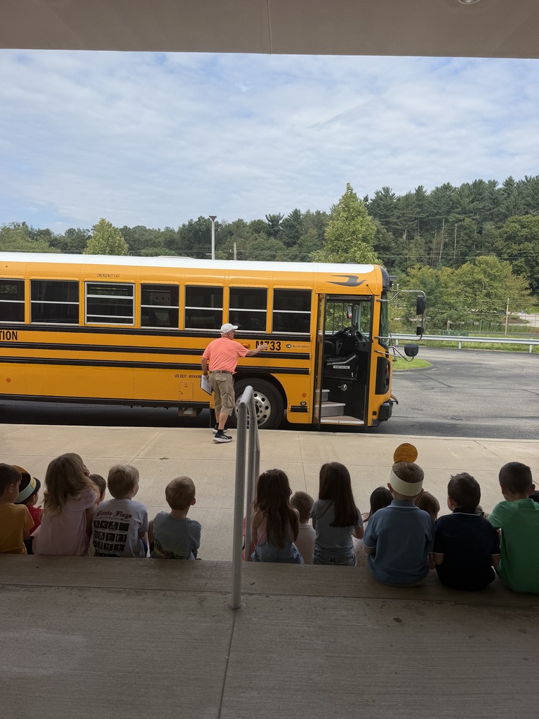 A school bus stands open-doored in a parking lot, with children seated on the curb. A person stands at the bus entrance. Trees and a streetlight are in the background.