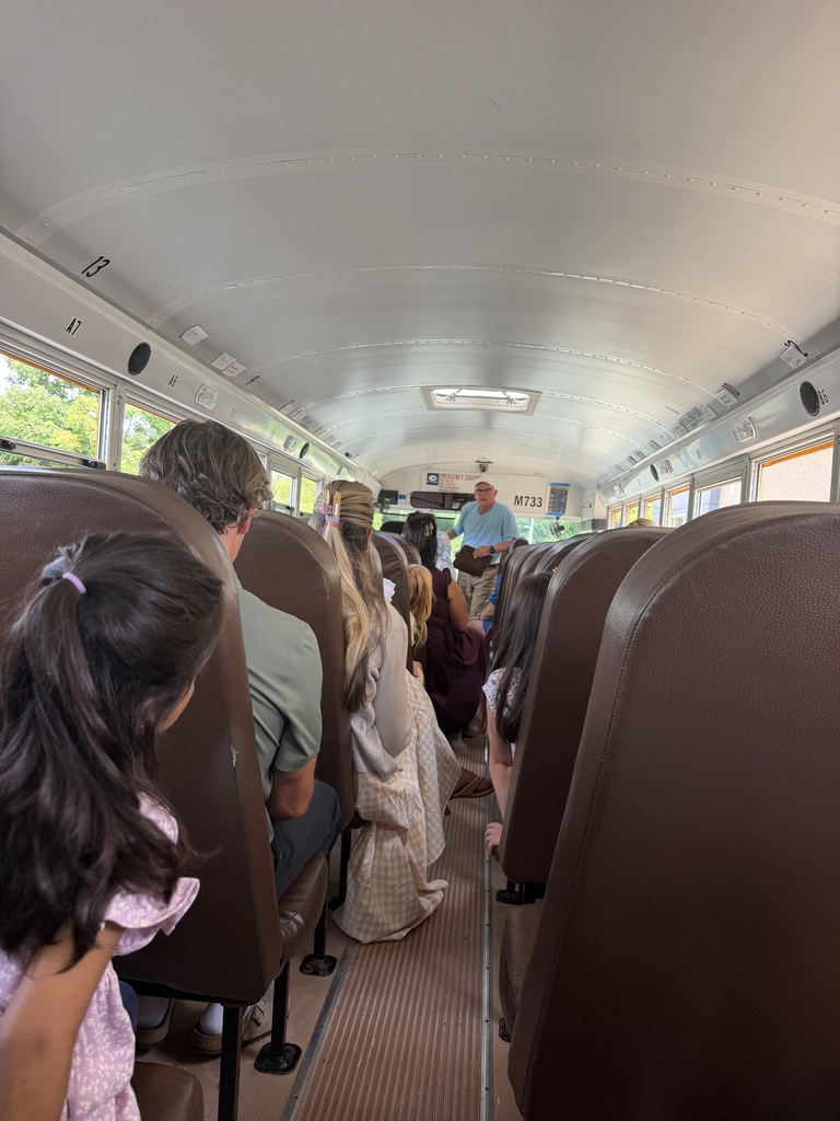 People sit in seats on a school bus. A person stands at the front. The bus has a white ceiling.