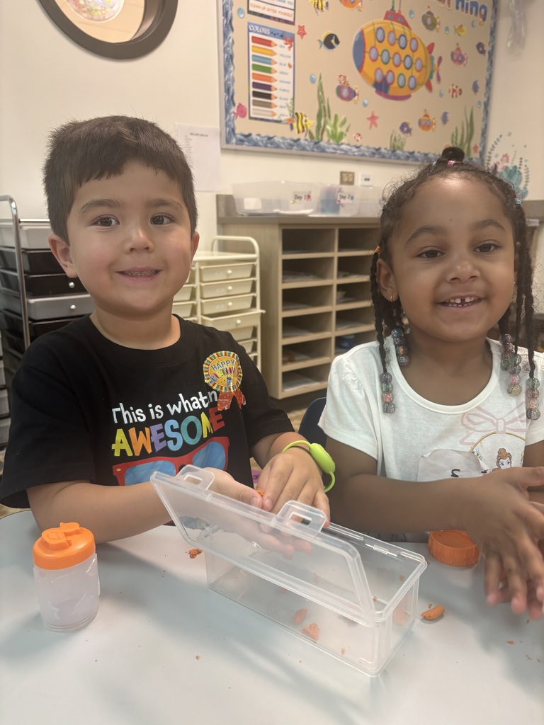 Two kids sitting at a table, one wearing a black t-shirt, the other in a white shirt. They are playing with objects in a plastic container.