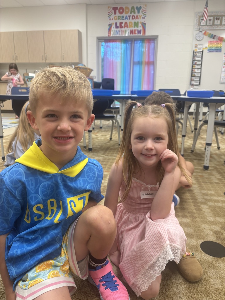 A young boy and girl are sitting on the floor. He wears a blue shirt and shorts, she a pink dress. Behind them, desks and chairs are arranged in a classroom.