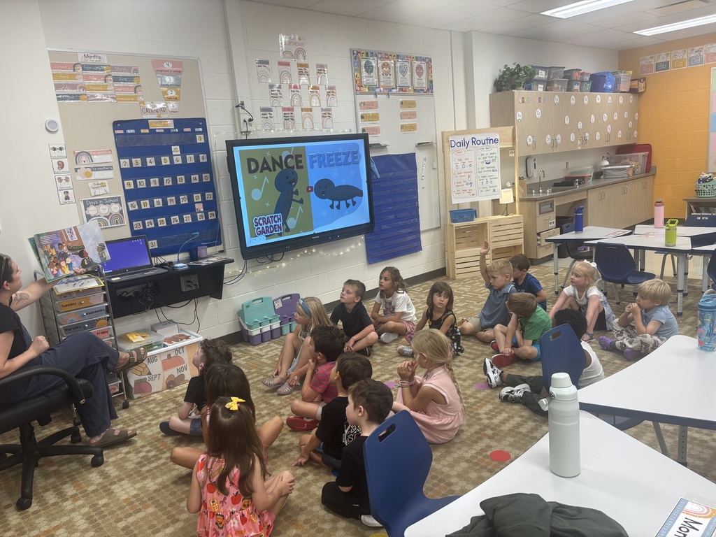 Kids sit in a circle on the floor in front of a teacher. A screen displays the word "Dance" above a frozen screen.