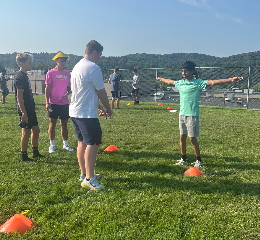 Five people on a field. One person in green wears a hat. They hold their arms out. Orange cones are on the grass.
