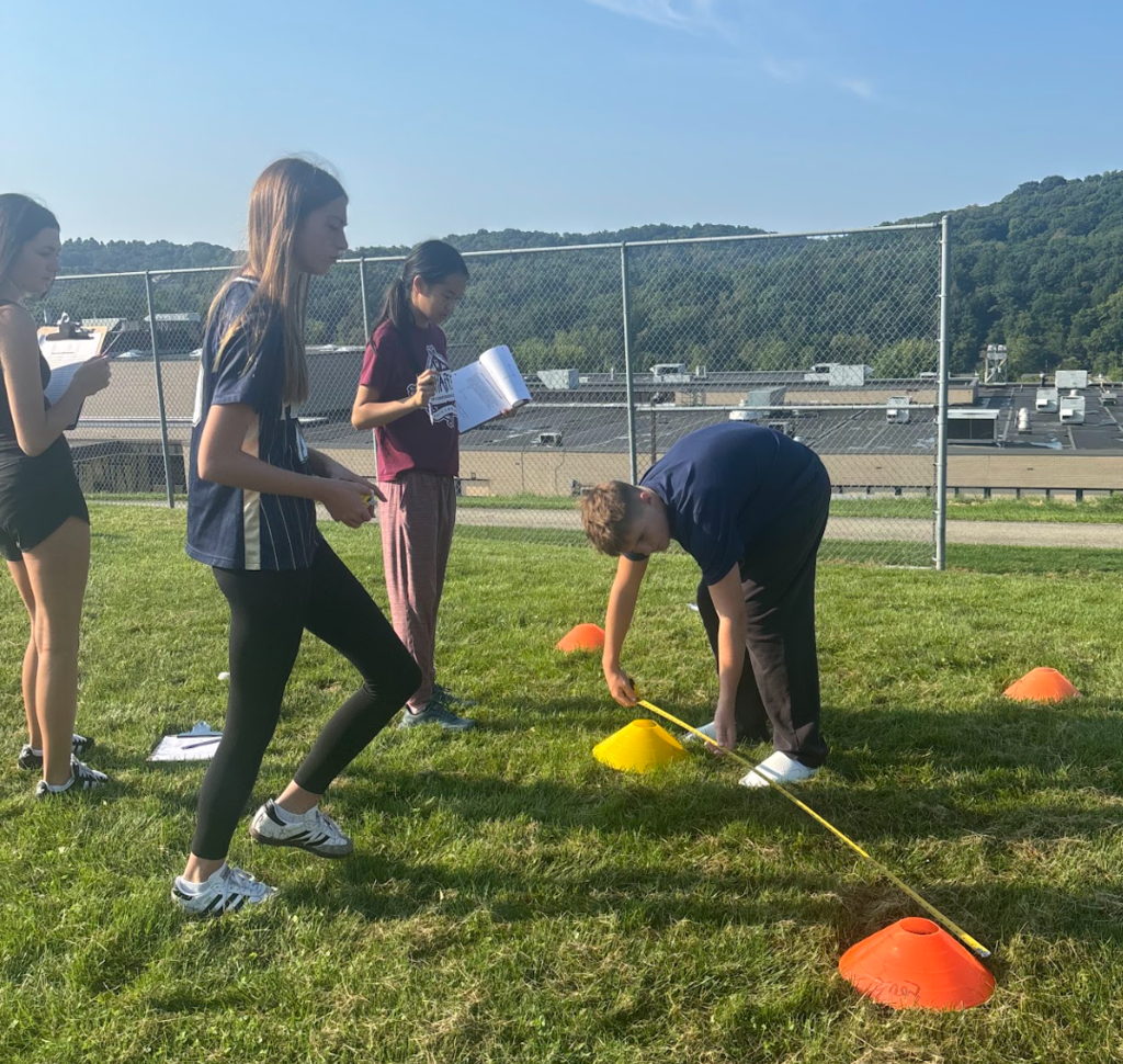 Four individuals on a grassy field. One person crouches with a measuring tape. Two hold papers. Colorful cones on the ground.