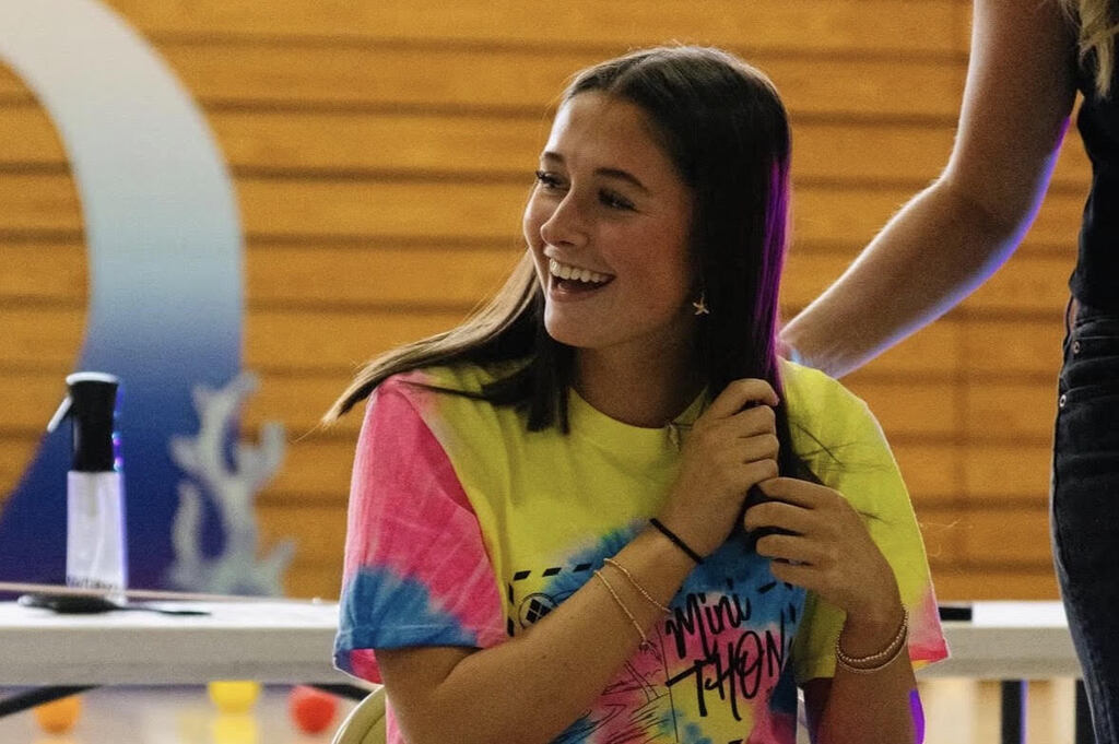 Regan Lynch with a tie-dye shirt smiles while another person styles her hair, with a table and blue wave design behind them.