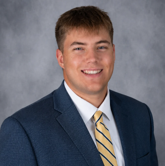 Person in navy suit and yellow striped tie standing against a gray background, smiling.