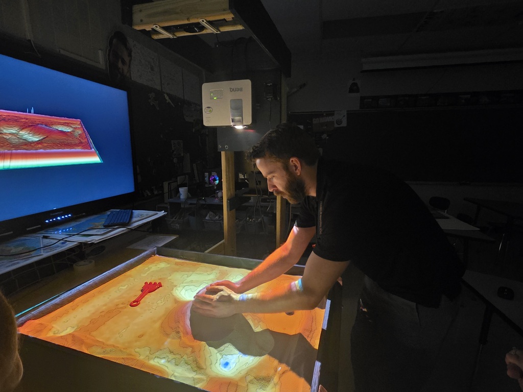 Man with a black shirt working on a lighted sand table with a large screen above.