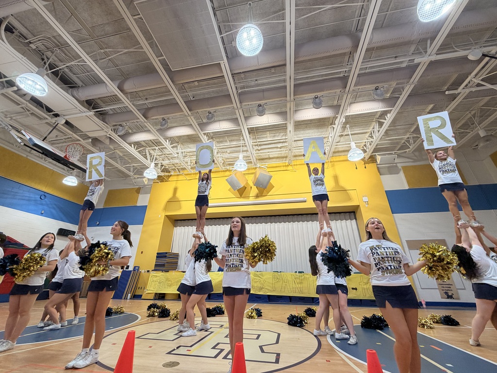 A group of cheerleaders in white tops and blue shorts performs stunts in a gymnasium.