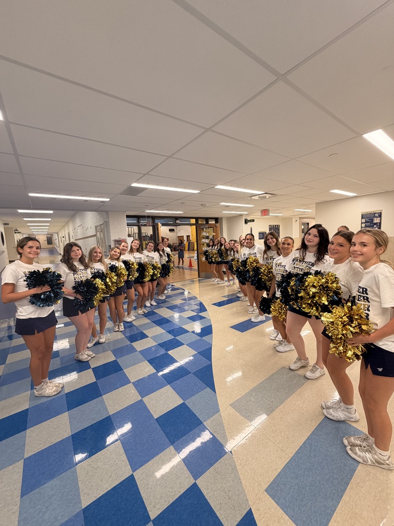 Group of cheerleaders in white shirts and black shorts holding pom-poms, standing in a hallway with a blue and white checkered floor and white ceiling lights.