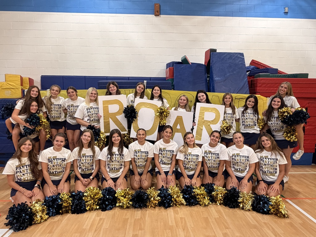A cheerleading team poses for a photo, holding a sign that reads "ROAR" in a gymnasium with colorful mats and equipment in the background.