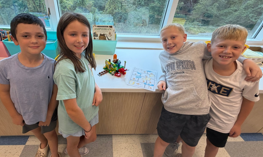 Four children pose in front of a desk. One holds a map and building blocks. A window shows trees.