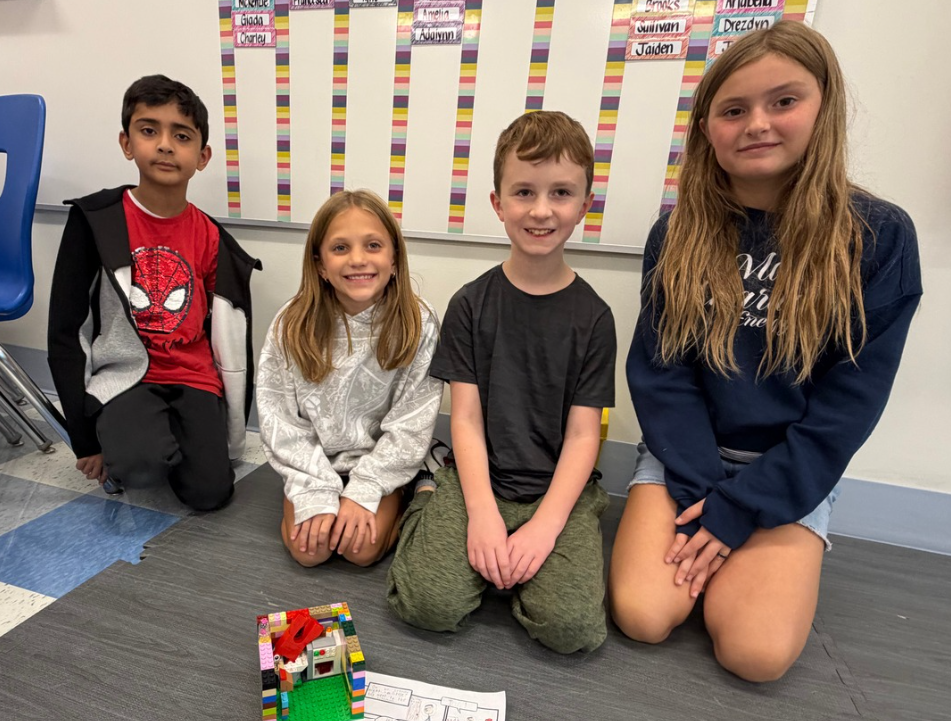 Four children seated on a carpeted floor, smiling. They pose for a picture, with one child holding a Lego creation.