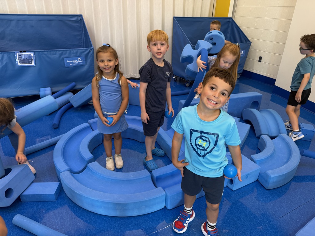 Children are playing in an indoor play area with blue mats and foam shapes. Some wear casual clothing, and others wear glasses.