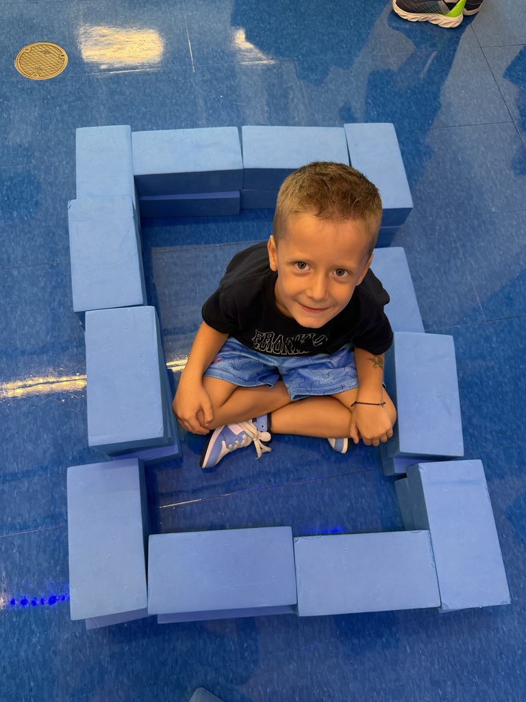 A young boy sits in a square formed by blue foam blocks, smiling and looking forward.