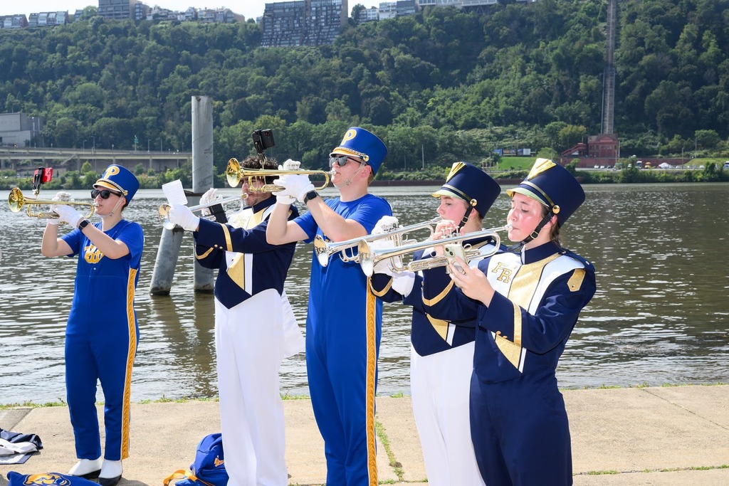 Five people in blue uniforms play trumpets on a river's edge, with a green hillside and trees in the background.