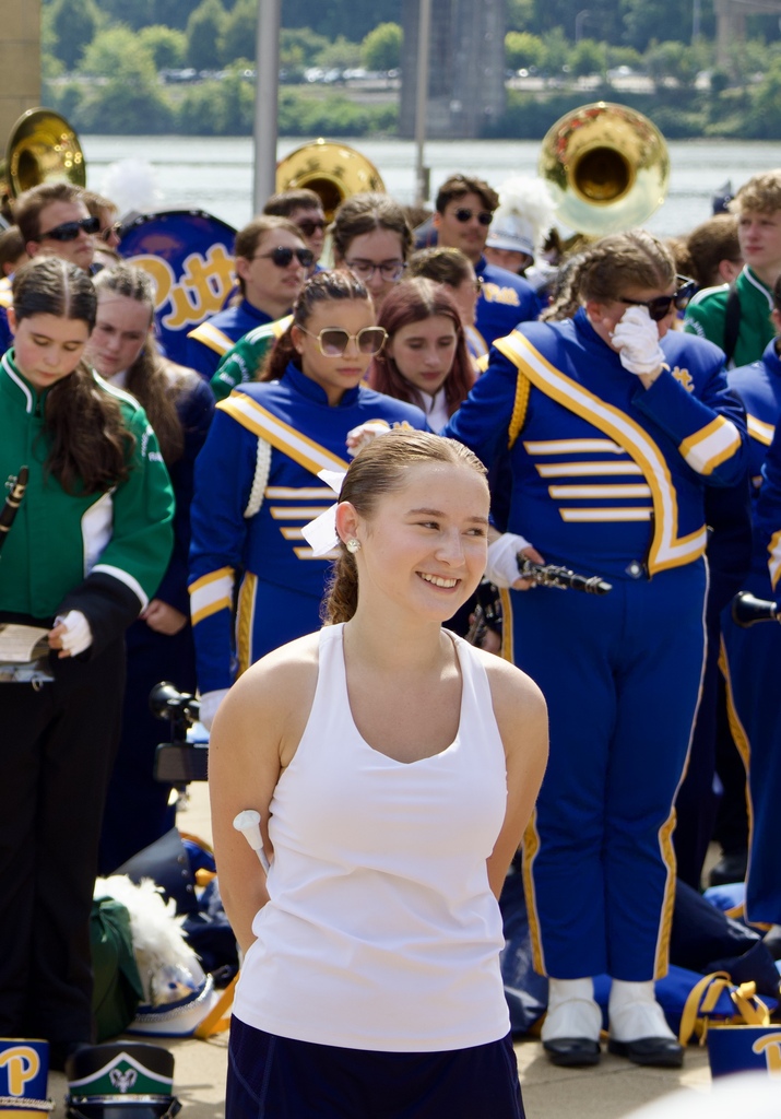 A girl in white stands in front of a marching band in blue and gold uniforms, near a river and bridge.