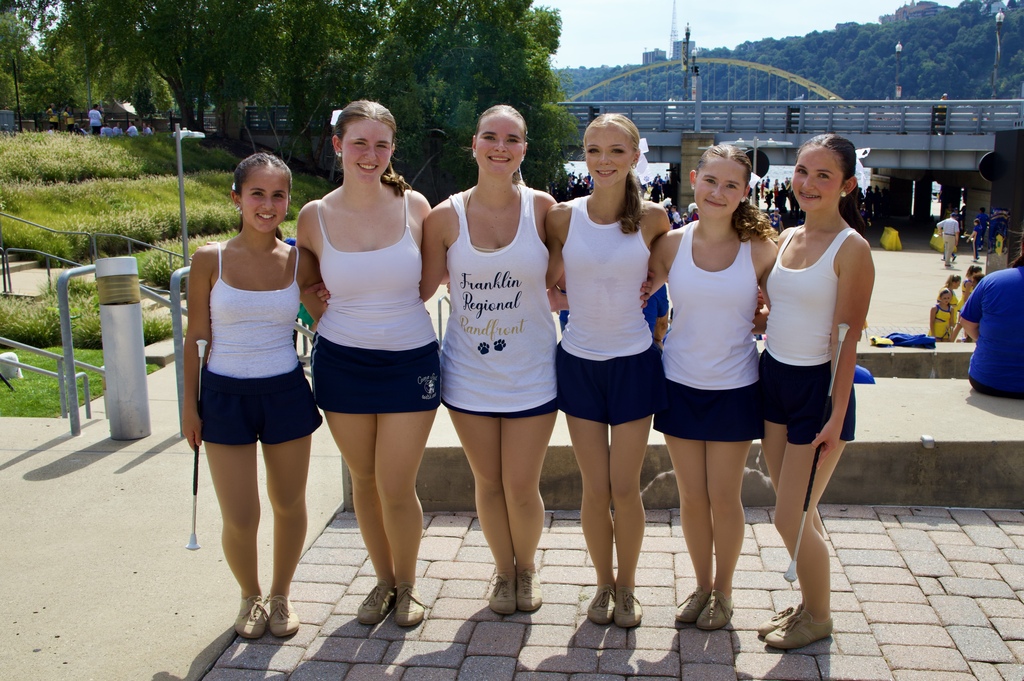 Five women in white shirts and blue shorts stand together on a brick path. Behind them, there's a bridge and a grassy area.