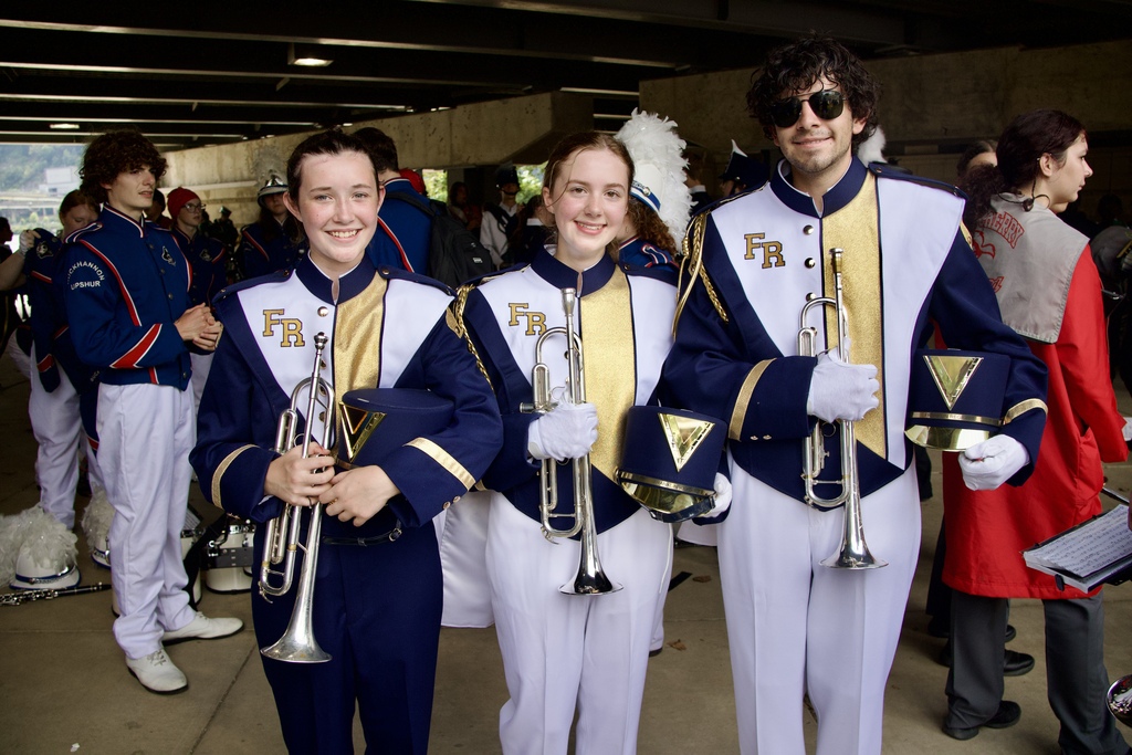 Three musicians in matching uniforms hold trumpets. They stand in a group of others in a tunnel.