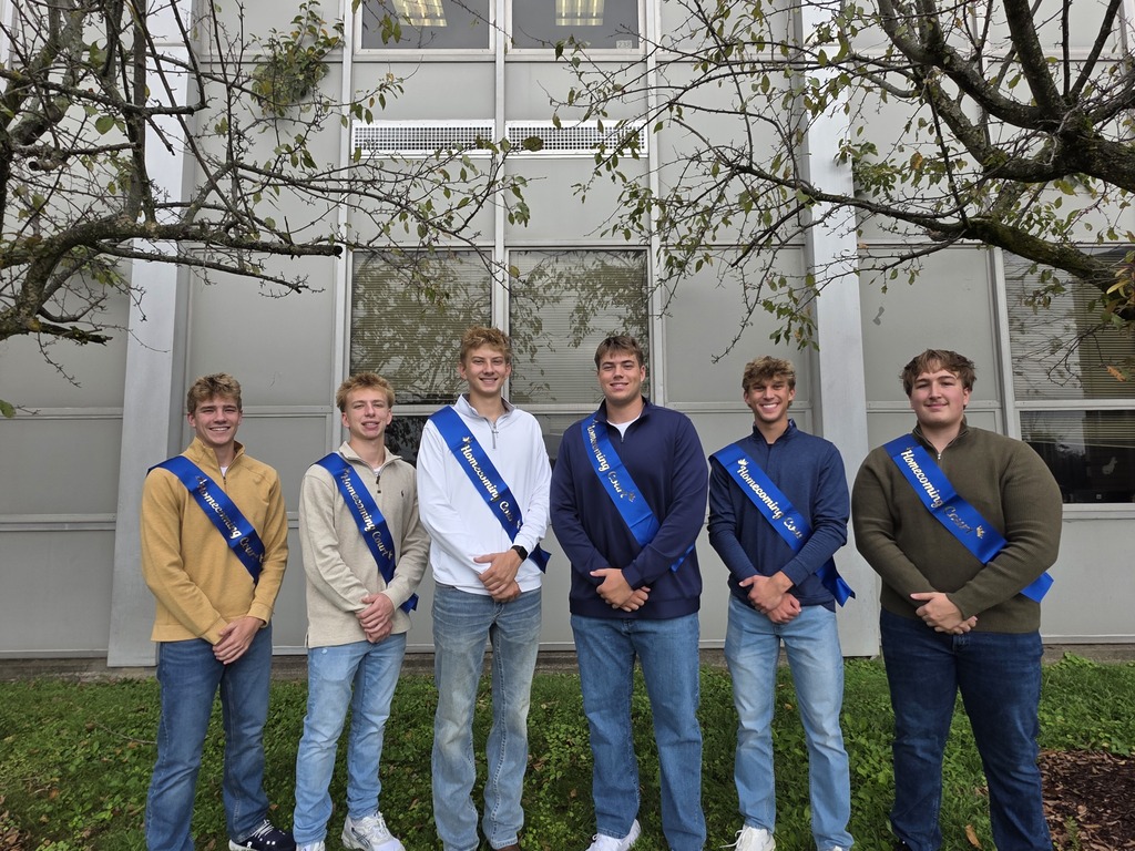 Six people wear sashes in front of a building. They stand on grass. Two trees are behind them.