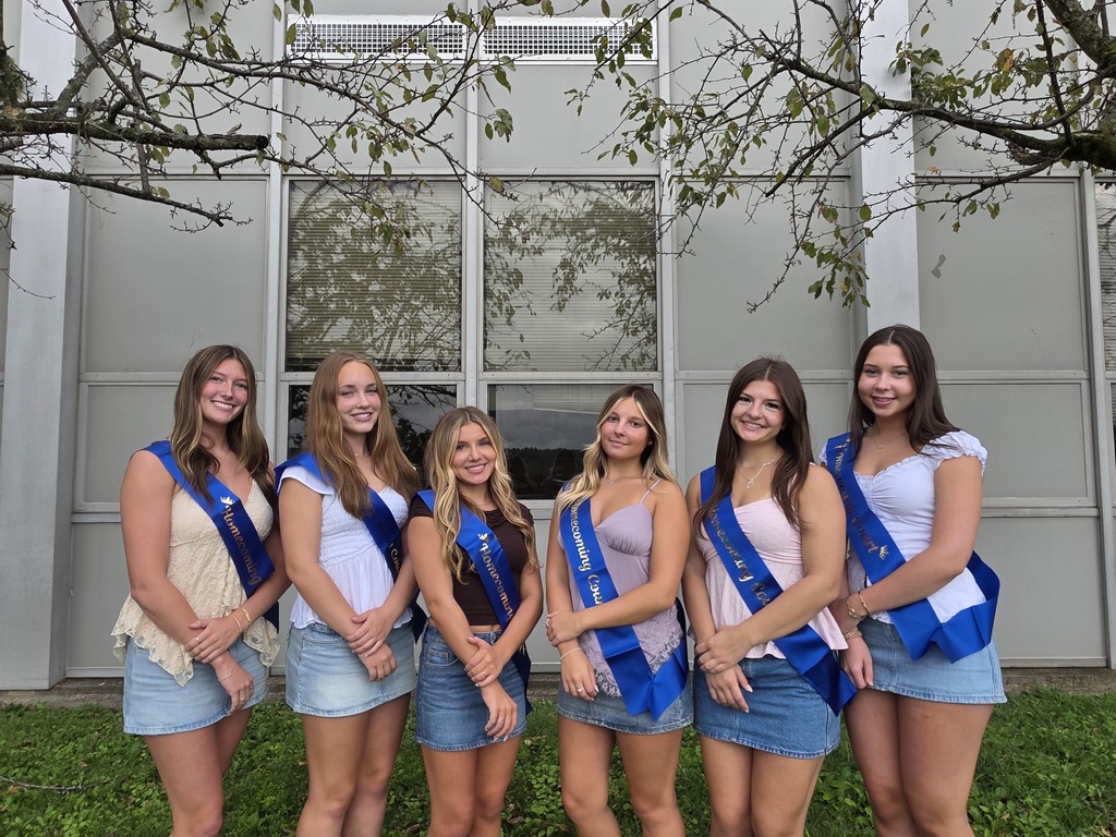 Six young women in sashes and denim skirts stand in front of a modern building with large windows.