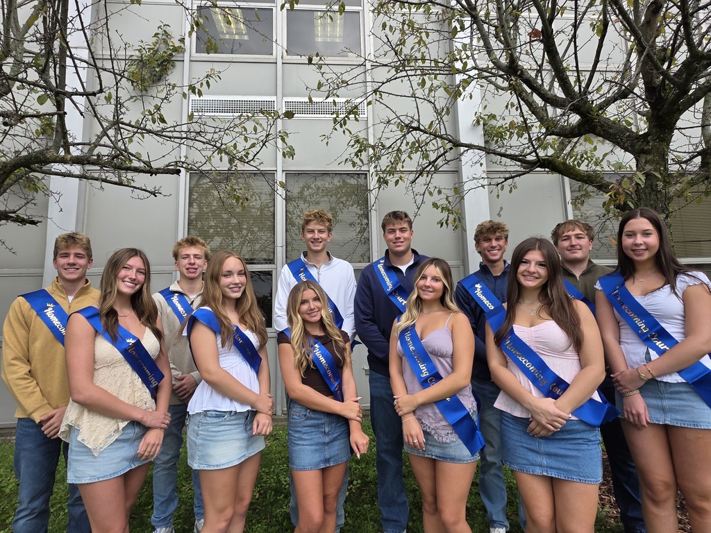 A group of people dressed in denim and blue sashes stand in front of a building with trees.