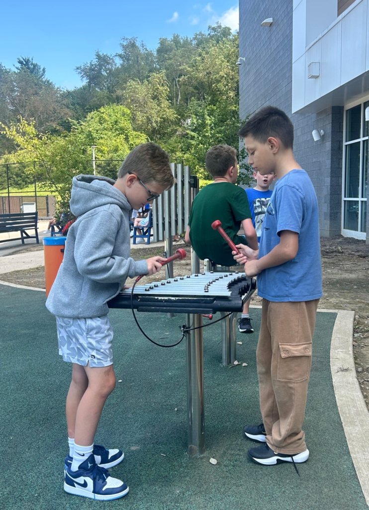 Three boys play xylophone in a playground. One holds mallets, another watches. Behind them, a building and trees.