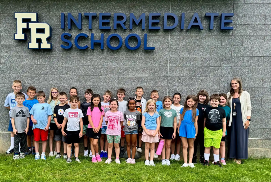 Group of students and a female teacher pose in front of the "FR Intermediate School" sign.