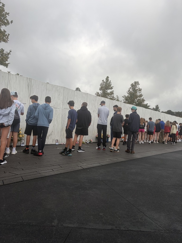 People in various attire stand in line before a white wall, with trees and a cloudy sky in background.