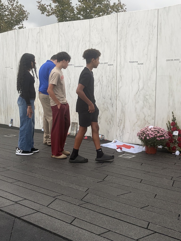 Five individuals stand near a white marble wall with engraved names. A bouquet of pink flowers lies on the ground.