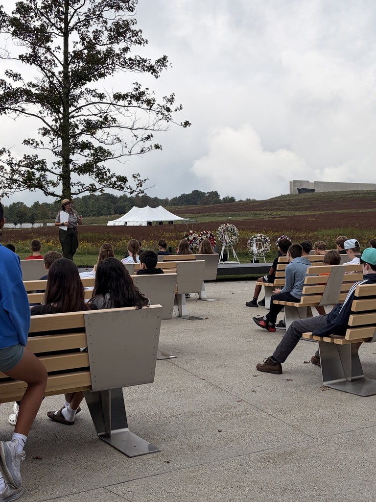 A group of people sit on benches around an outdoor amphitheater with a speaker. A tree stands in the background.