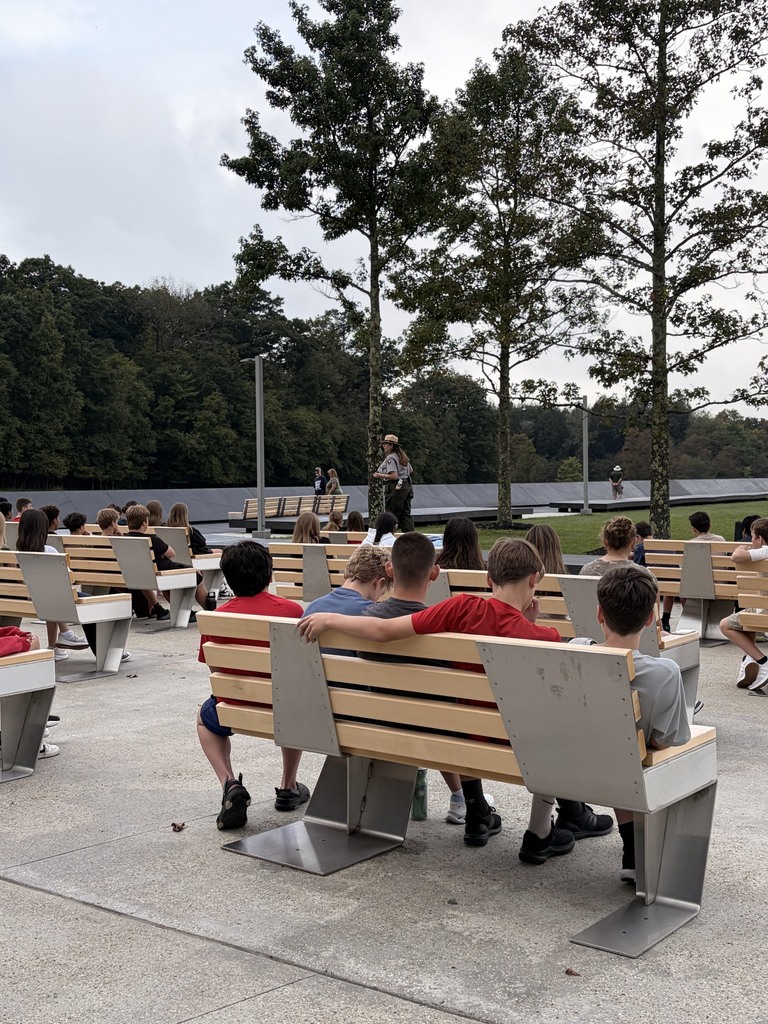 A group of children sit on benches in an outdoor area, surrounded by trees. A person stands in the background.