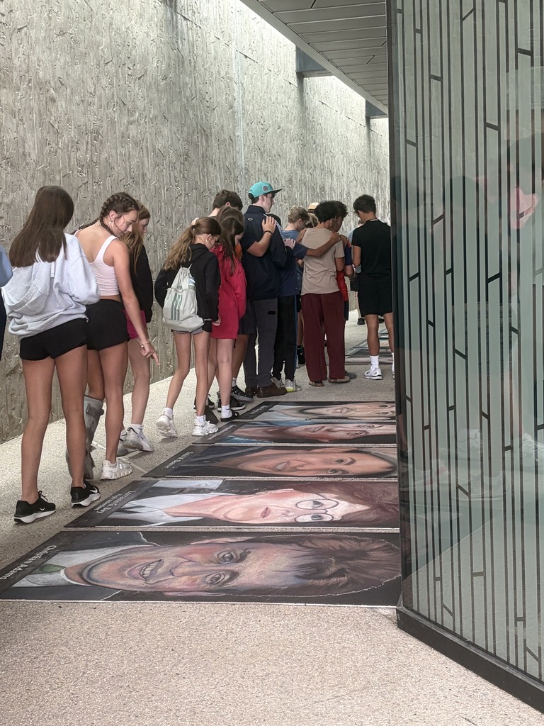 Visitors stand in a line at a museum, looking at portraits on the floor. The portraits depict a person in various poses.