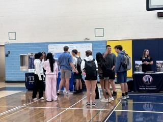 A group of people wearing casual clothing and backpacks stand in a gym with a blue floor.