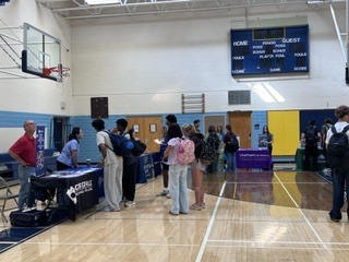 A gymnasium with people standing and talking, a table with a sign, and a scoreboard in the background.