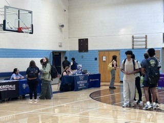 People gather around tables in a gym. Tables have blue covers. One table has a sign reading "WESTFIELD HIGH."