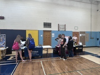 Group of people in a gym. Some are standing at tables with signs. There are blue walls and wooden floors.