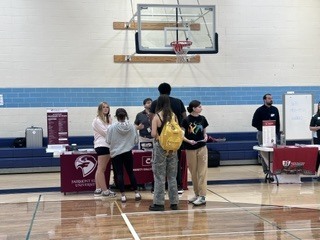 People stand at tables with promotional materials in a gym, with a basketball hoop in the background.