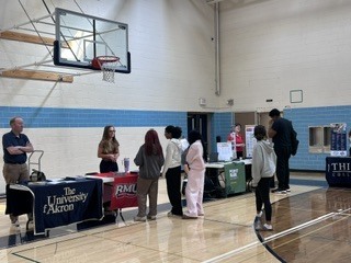 People stand at tables with banners in a gymnasium. A basketball hoop is visible in the background.