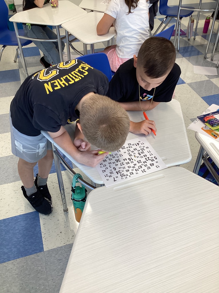Two students work on a paper with a puzzle grid in a classroom. A third student sits in the background.