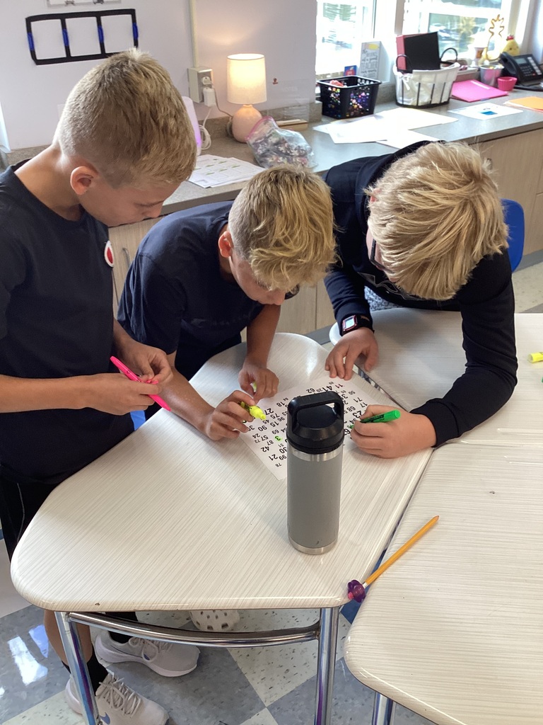 Three boys in a classroom, seated at a table. They are working on a puzzle with a variety of colored markers.