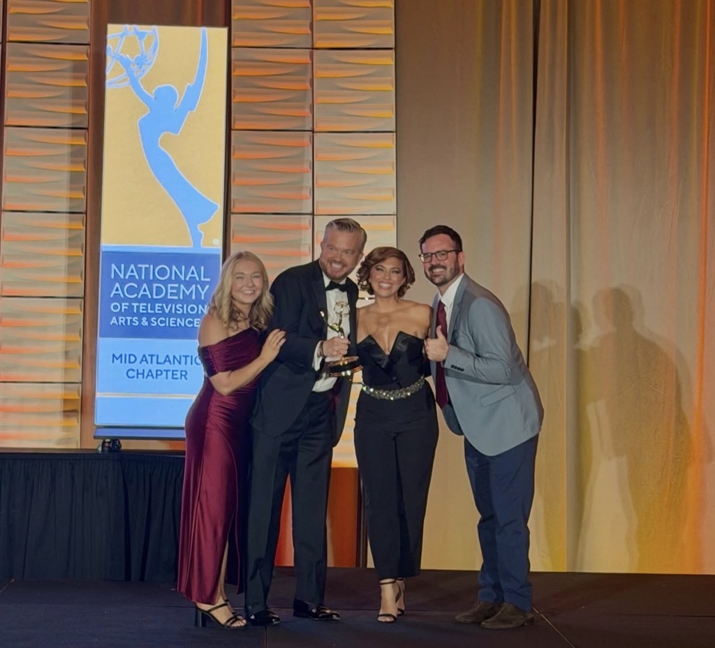 Four people stand on a stage with a sign behind them that reads "National Academy of Television Arts and Sciences Mid Atlantic Chapter."