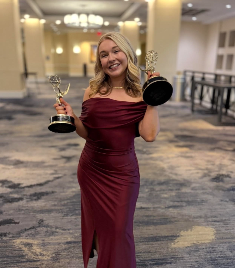 A smiling woman holds two awards in a hallway with marble flooring. She wears a maroon dress.