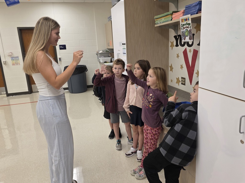 Woman standing, speaking to a row of children. Children in various outfits, standing and crouching. Background: hall with lockers and doors.