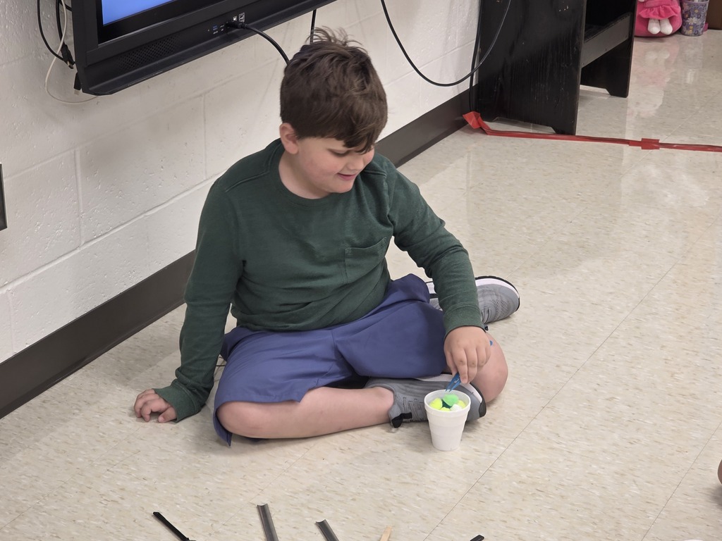 A child in a green long sleeve and blue shorts sits on the floor holding a cup.