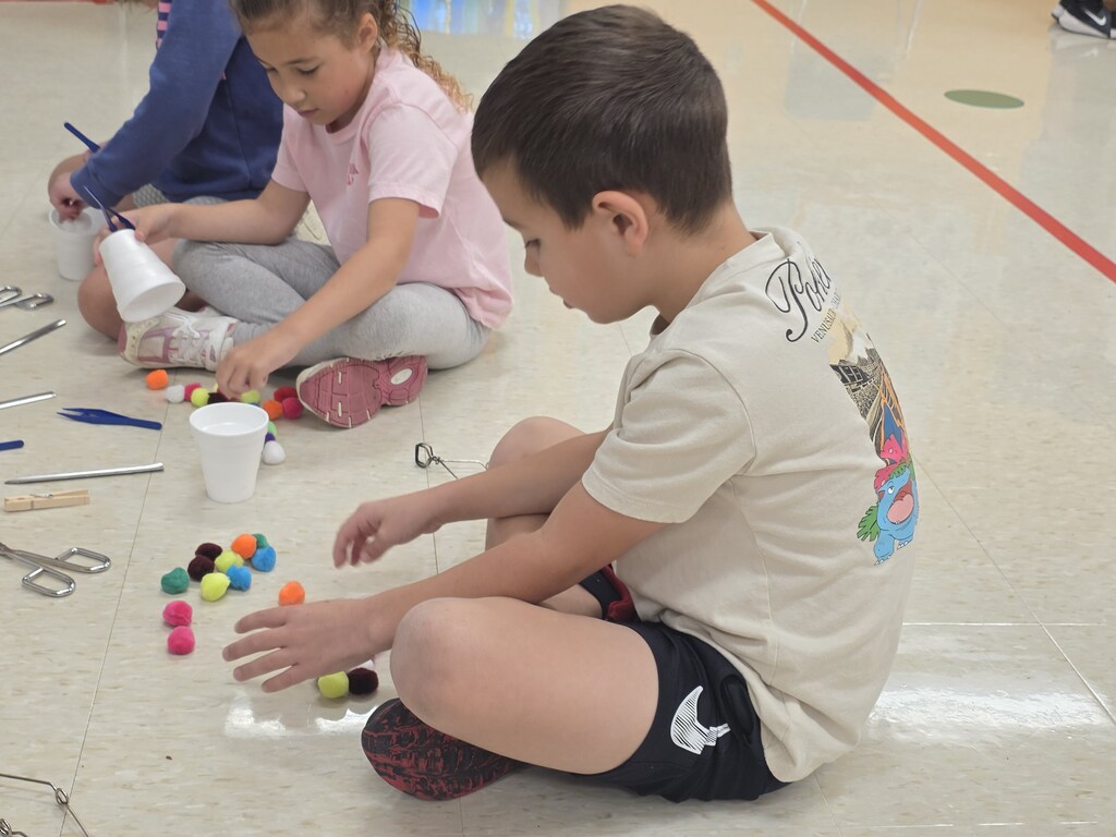 Two children are on the floor, playing with colorful balls. One child uses a cup. They sit on a white floor with a red line.