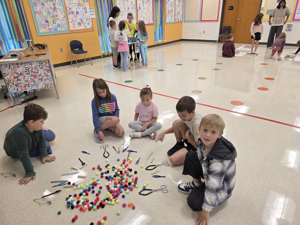 Kids sit on floor, creating a circle with colorful beads. Background shows a room with tables, chairs, and children.