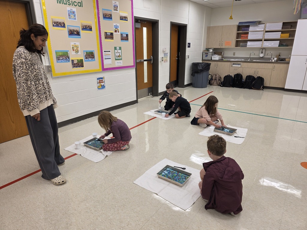 An adult woman stands by a bulletin board while children sit in a classroom, each using a tablet on paper.