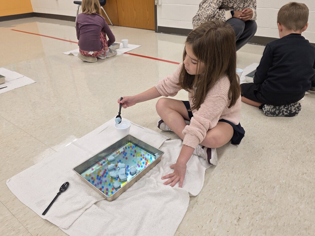 Children seated on the floor, one mixing colorful paint on a tray with a spoon, others observing.